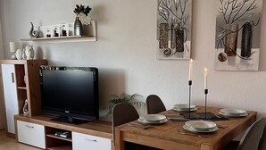 A modern dining area with a wooden table set for two, a flat-screen TV on a wooden console, and two wall-mounted paintings.