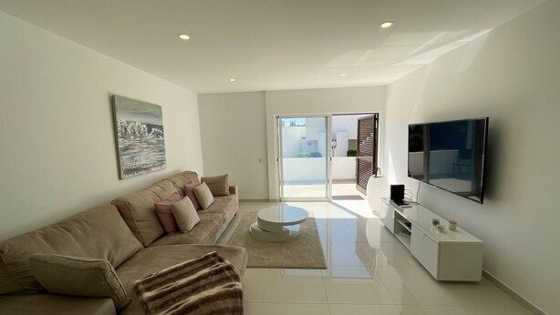 A modern living room with a sofa, a coffee table, a flat-screen TV, and a sliding glass door leading to a balcony. - Casa Rosa - Vilamoura (Vilamoura)