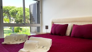 A neatly made bed with a red bedspread and two white towels with a logo, a window with blinds, and a view of greenery outside. - Casa Rosa - Vilamoura (Vilamoura)