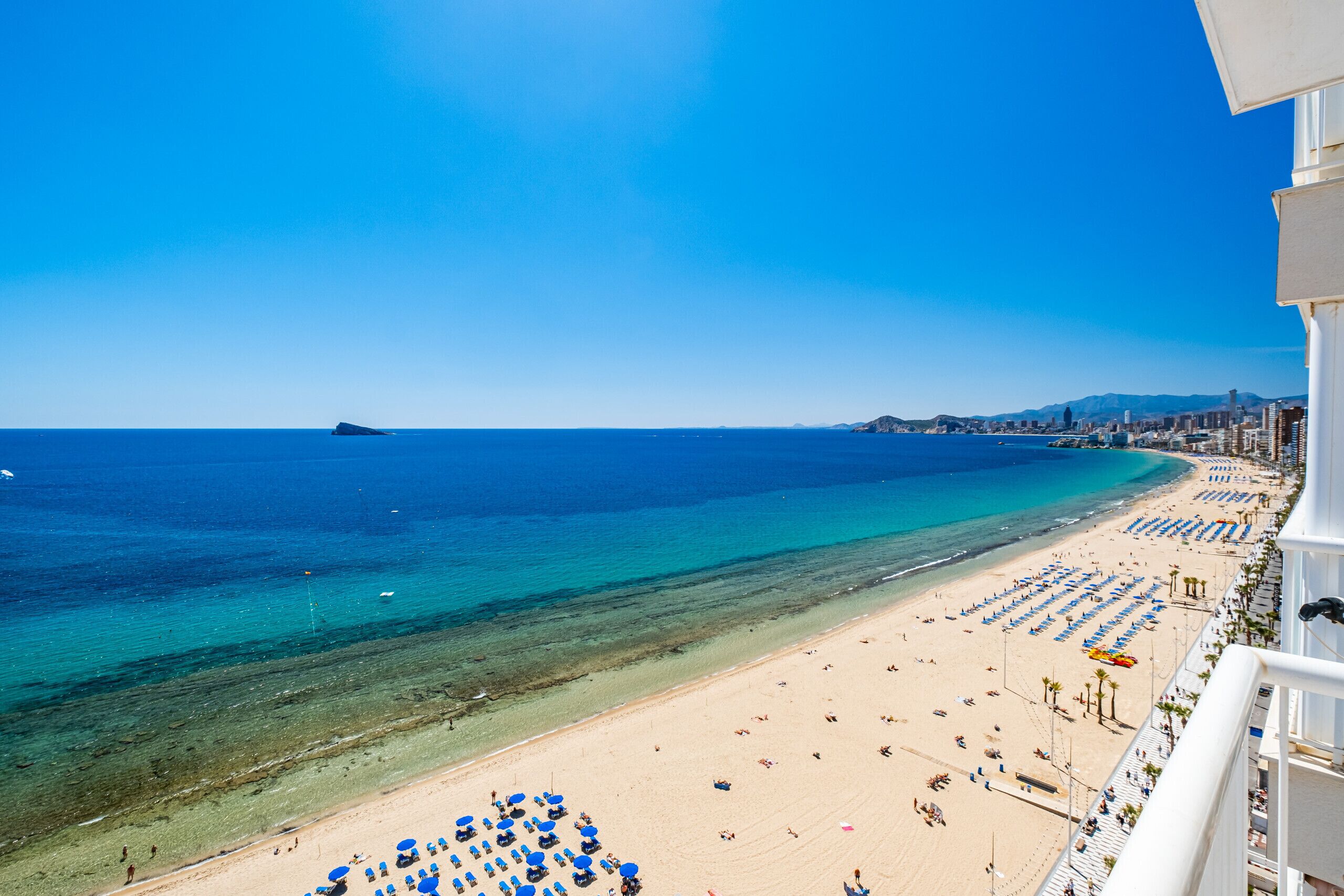 Una playa con hileras de sombrillas azules y tumbonas, un mar azul cristalino y un paisaje urbano al fondo.