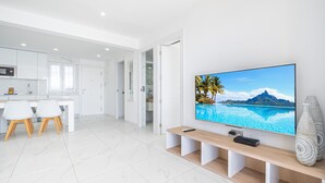 A modern kitchen with white cabinets, a dining area with a white table and chairs, and a large flat-screen TV mounted on a wooden console.