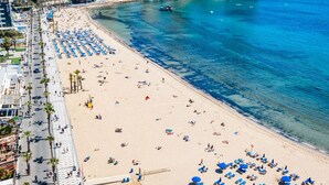 A beach with rows of blue umbrellas and sun loungers, a promenade with palm trees, and buildings in the background.