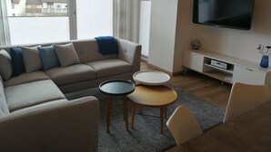 A modern living room with a sofa, two side tables, a TV, and a view of buildings through large windows. - Ferienwohnung Wiesenblume UR (Beilngries)