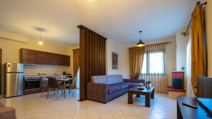 Una sala moderna con sofá, mesa de centro y mueble para televisión. Cuenta con una zona de cocina con refrigerador y estufa. La habitación tiene ventanas amplias con cortinas.