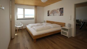 A bedroom with a wooden bed, a white chair, a bedside table, and a window with curtains. - Ferienwohnung Ranunkel (Beilngries)