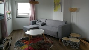 A modern living room with a grey sofa, a round coffee table, a colorful rug, and a window with pink curtains. - Ferienwohnung Ranunkel (Beilngries)