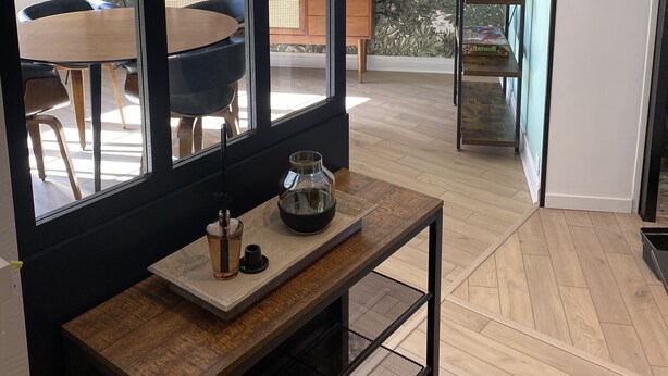 A modern interior with a large window, a wooden dining table, chairs, a small table with a vase, and a metal shelf with decorative items.