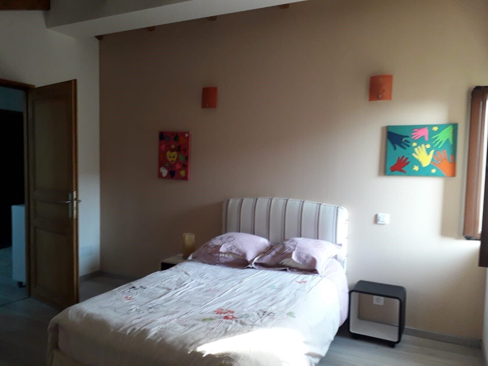Un dormitorio con una cama, almohadas, una mesita de noche, una pequeña mesa y dos lámparas fijadas a la pared.