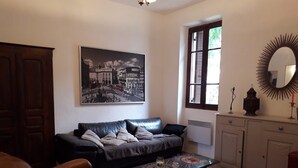 En stue med en sort lædersofa, et sofabord med en dekorativ midtpunkt, et stort bybillede i ramme, et vindue med skodder og et spejl med solstråle-design.