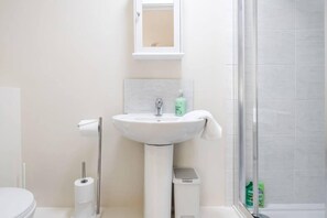 A bathroom with a white sink, a mirror, a toilet, and a shower area. - Cosy 2BR Maisonette Near Hospital & Stations (Bedford)