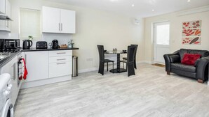 Een moderne keuken met witte kasten, een eethoek met een ronde tafel en stoelen, en een woonkamer met een zwarte leren bank en een rode kussen.