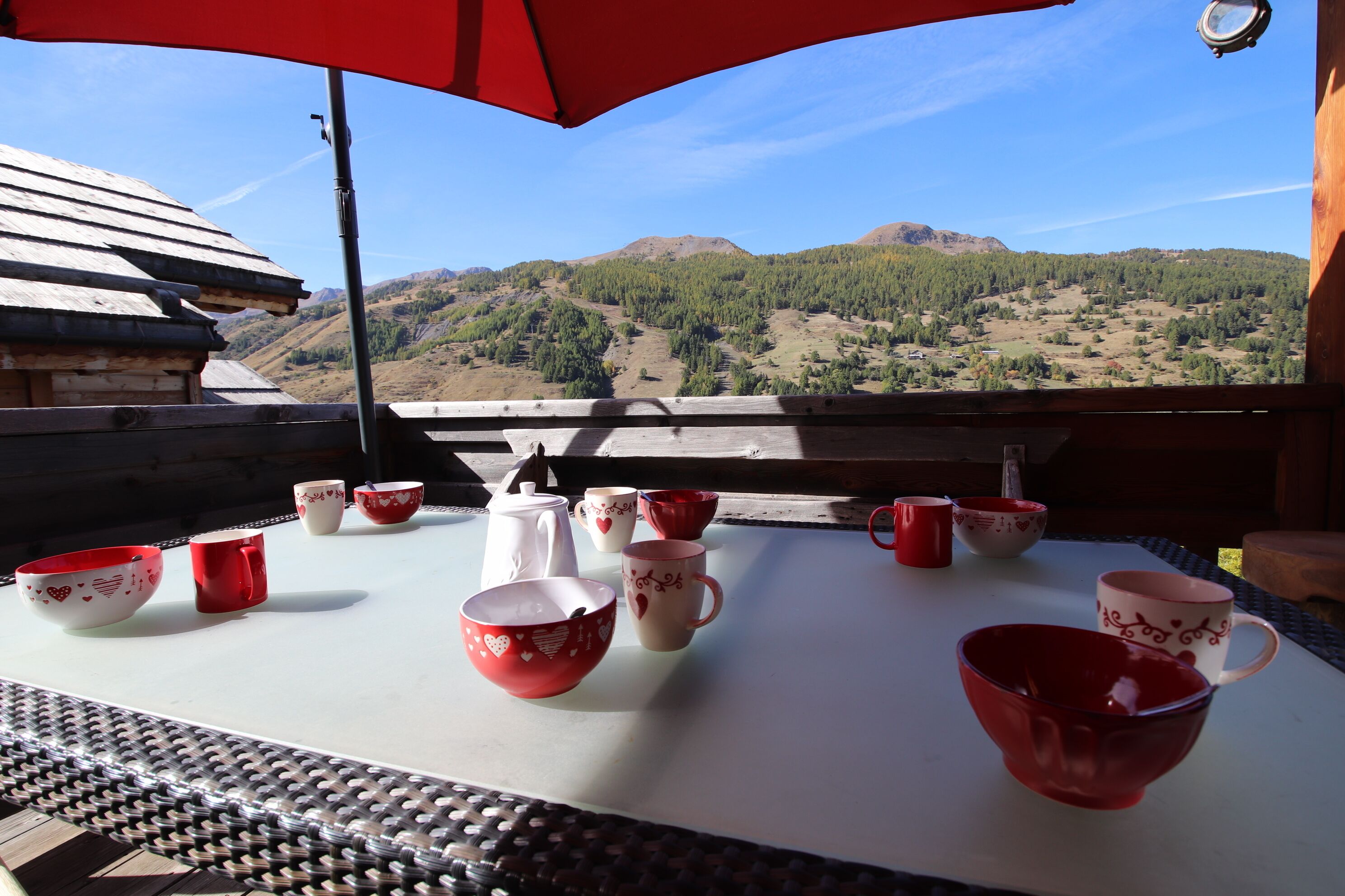 Taula amb diverses tasses de ceràmica vermelles i blanques i una gerra blanca, situada a l'aire lliure amb vista a les muntanyes i un cel serè.