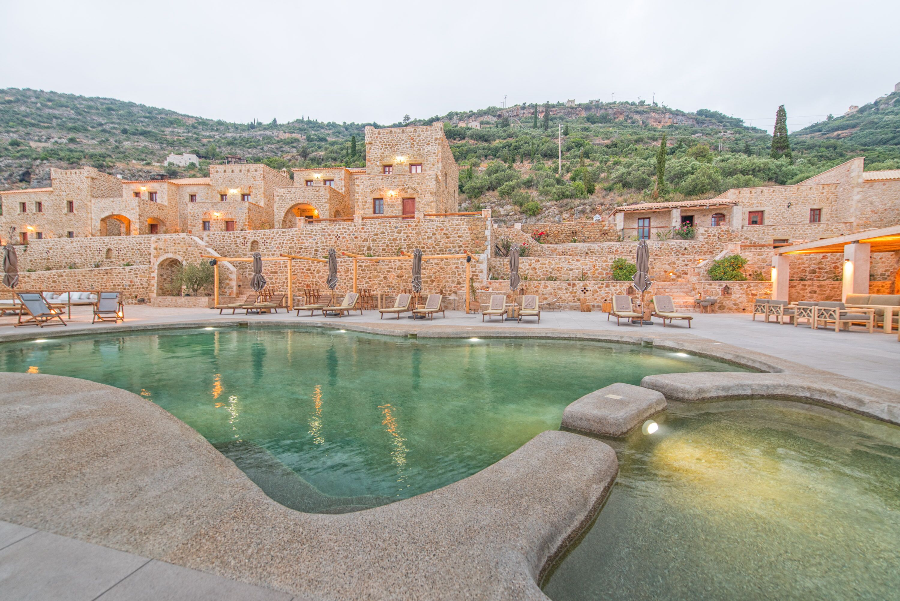 A stone pool with lounge chairs, surrounded by traditional stone buildings with arched doorways, set against a hillside backdrop.