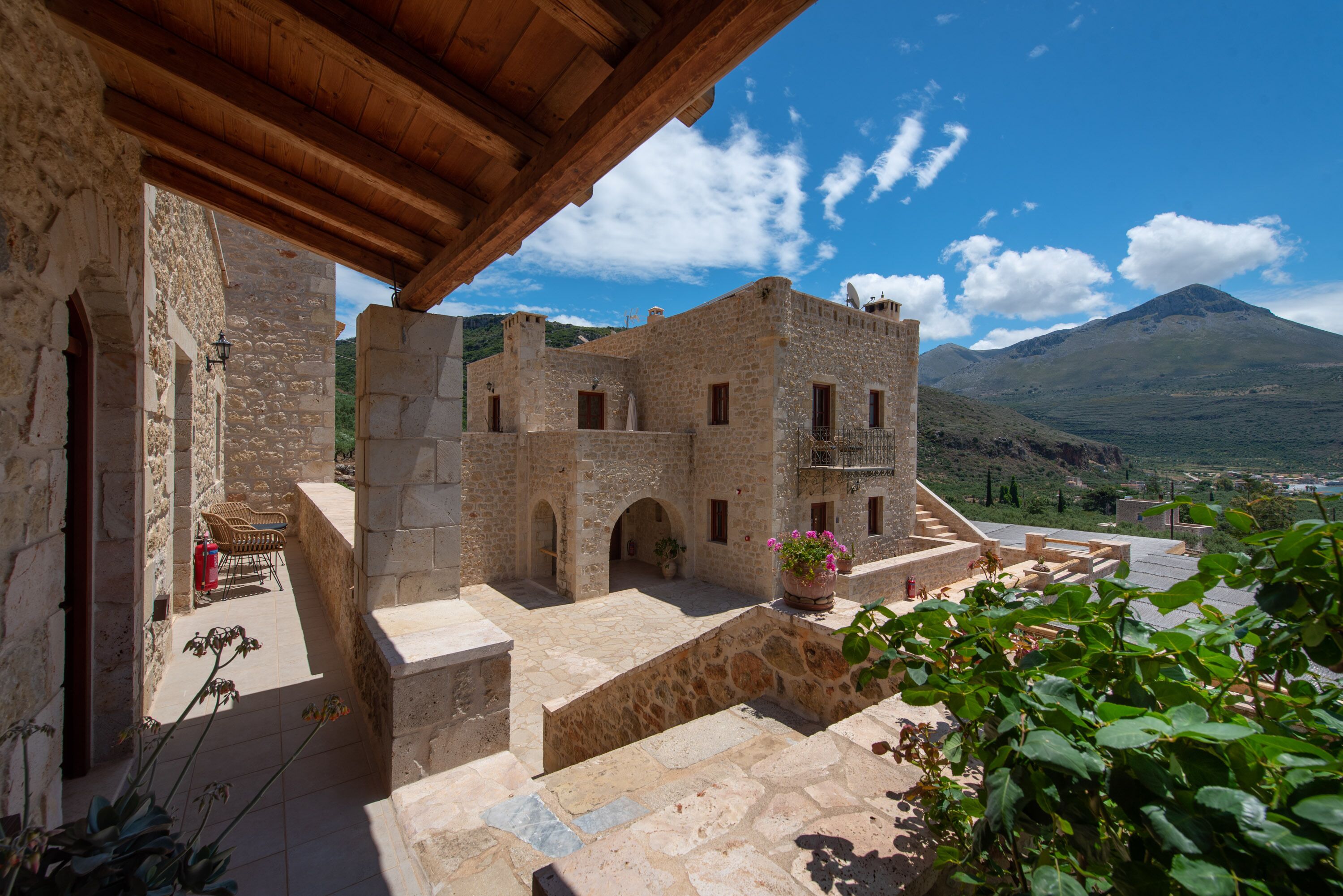 A stone building with a wooden roof, a courtyard with a bench, and a mountain in the background.