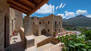 A stone building with a wooden roof, a courtyard with a bench, and a mountain in the background.