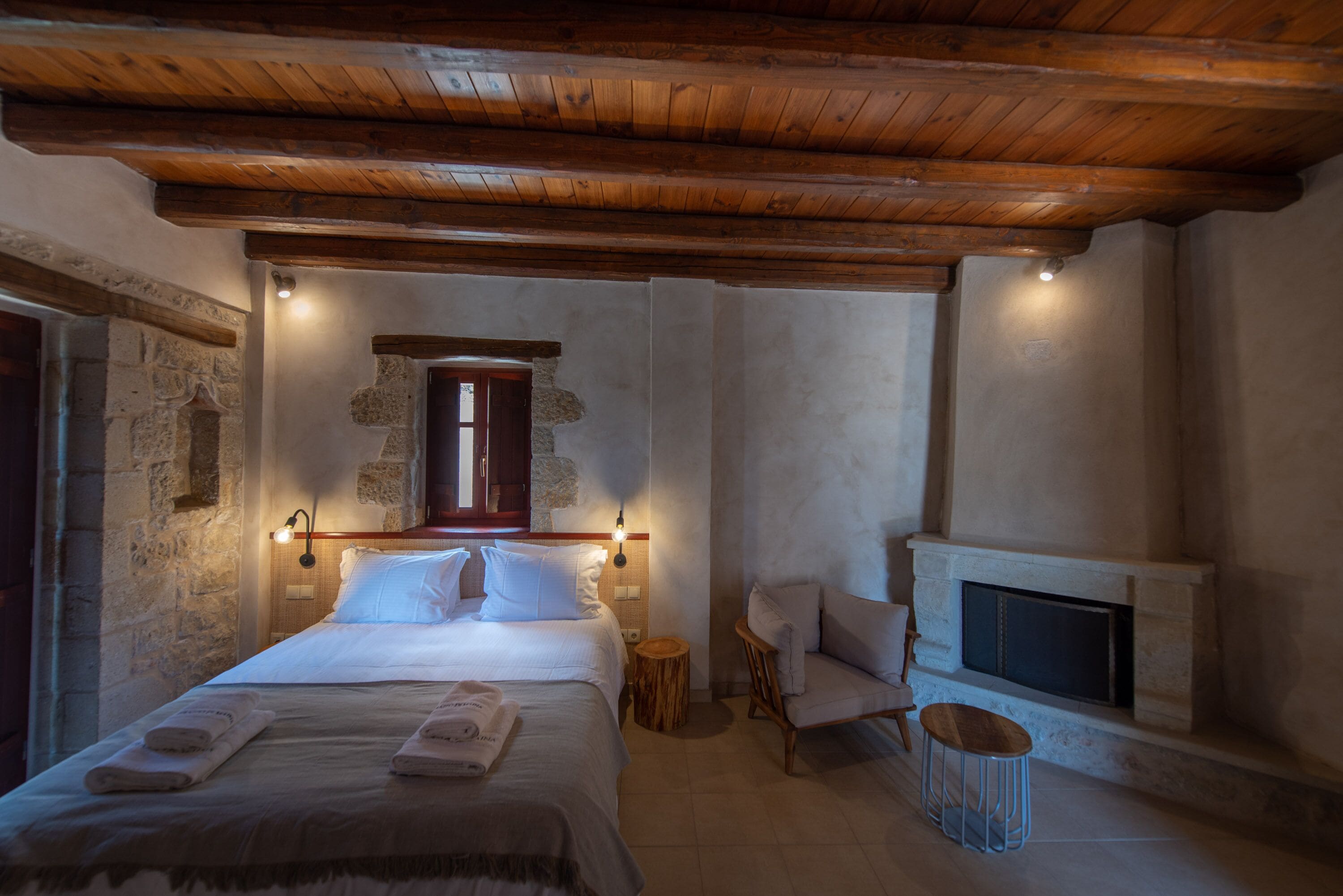 Een gezellige slaapkamer met een groot bed, een houten plafond, een stenen muur, een open haard en een kleine ronde tafel.