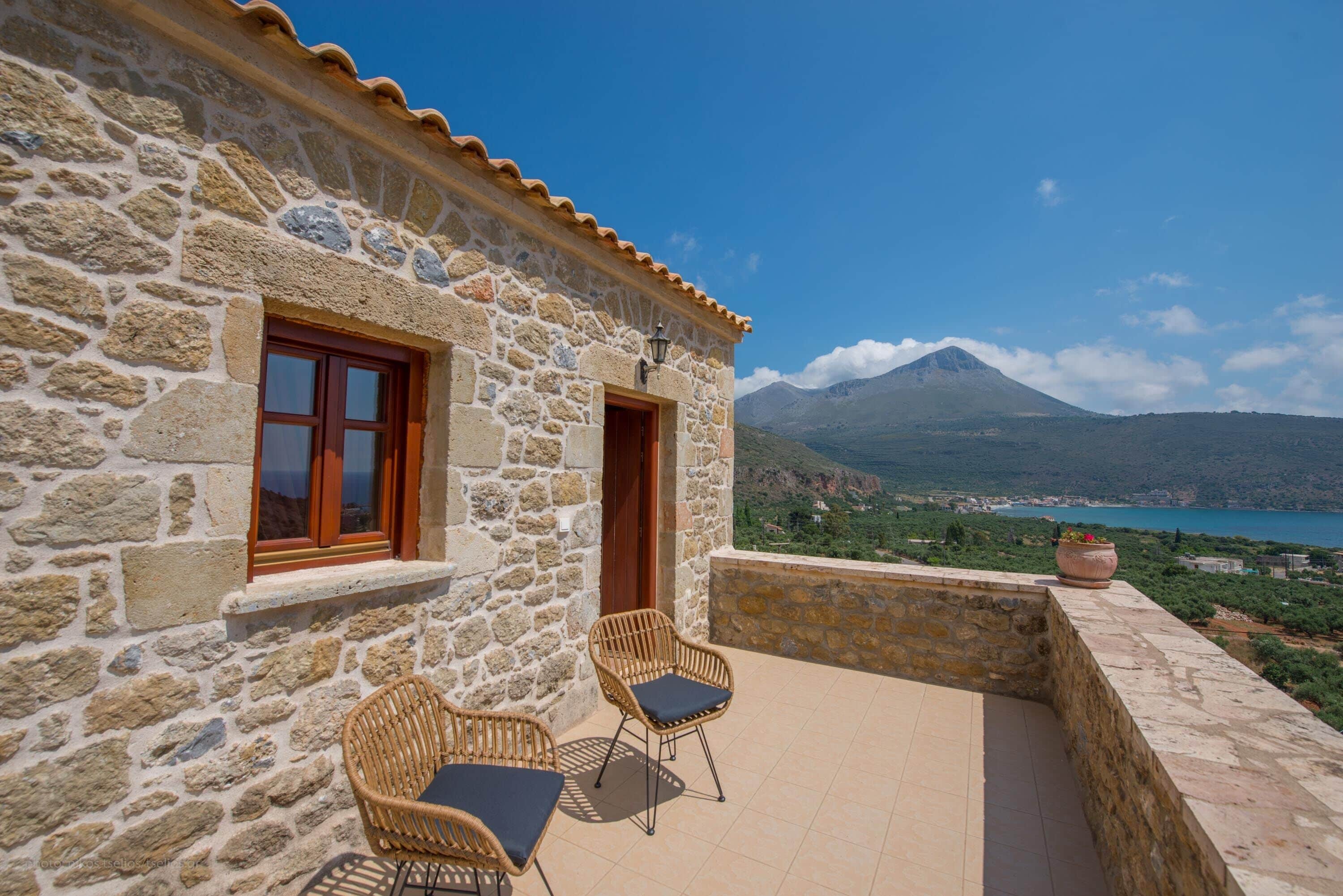 A stone building with two wicker chairs and a table on a balcony overlooking a mountainous landscape and a body of water.