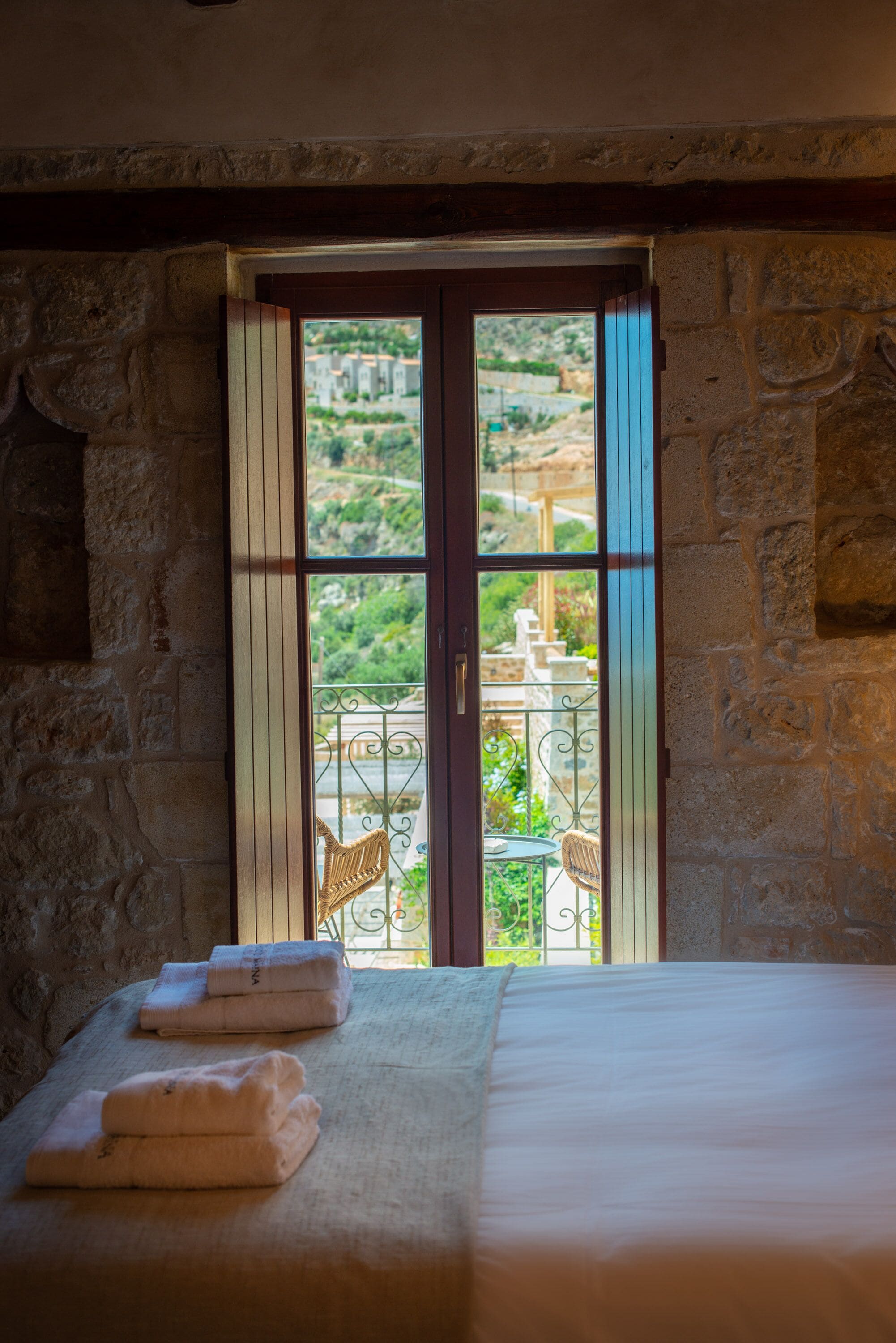 A room with a bed, a window with a view of a balcony, and a stone wall.