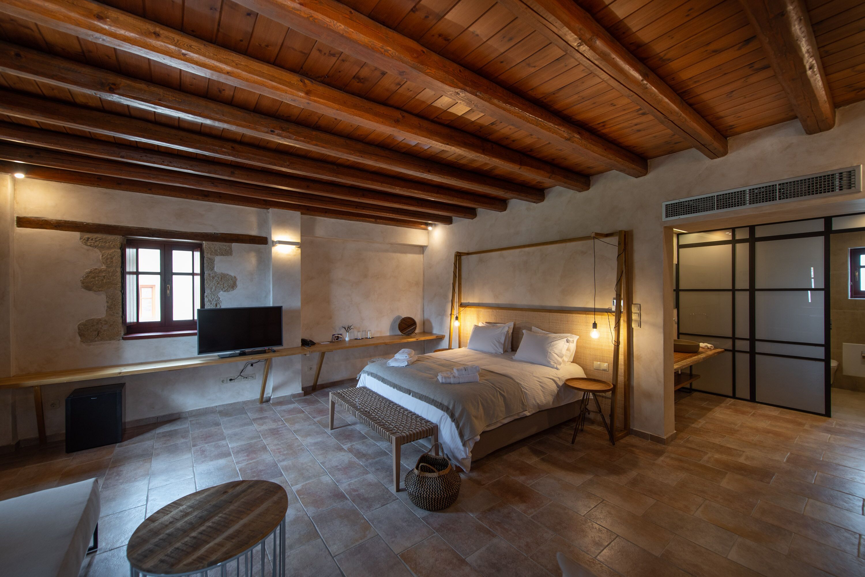 Un dormitorio con techo de madera, cama, televisor, banco y una pequeña mesa.