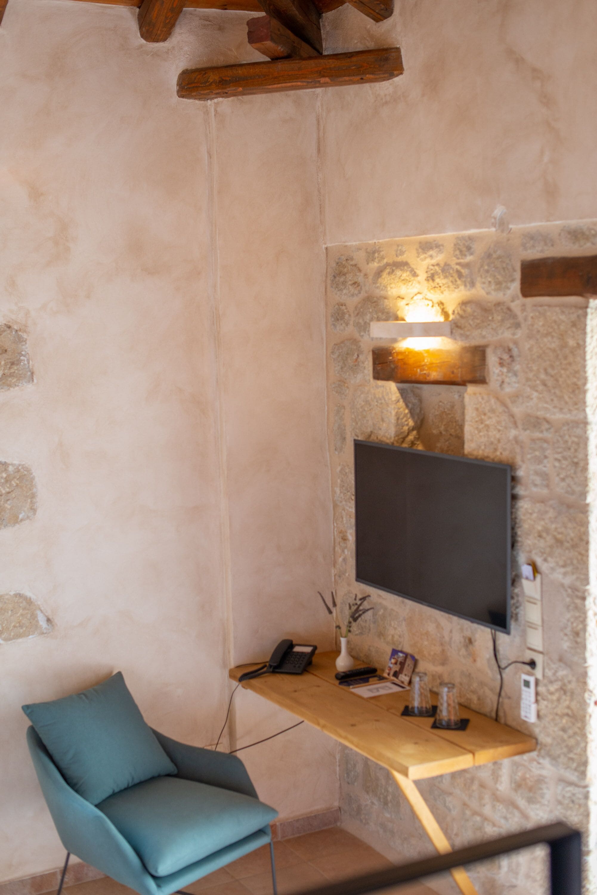 Une pièce avec un mur en pierre, un téléviseur à écran plat fixé au mur, un bureau en bois, une chaise de couleur turquoise et une petite table sur laquelle se trouvent un téléphone et quelques objets décoratifs.