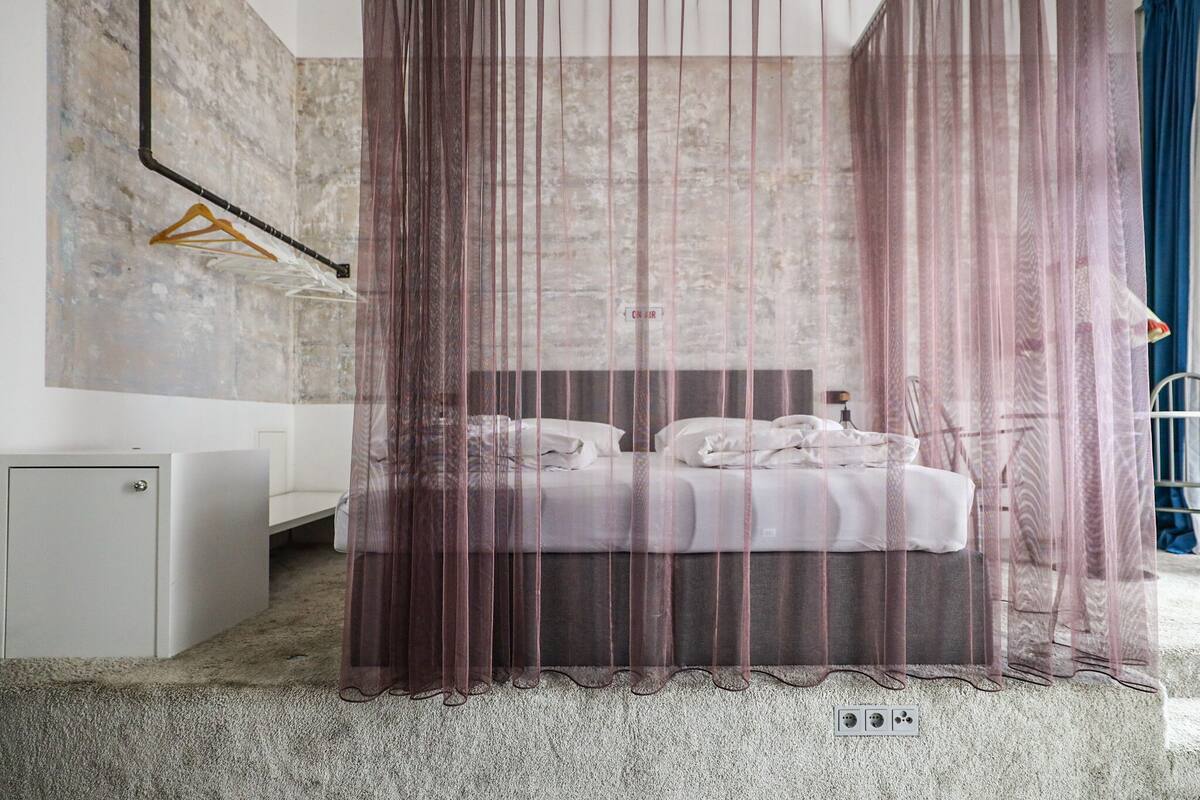 A bedroom with a bed, a bedside table, a hanging light, and a wall-mounted shelf.