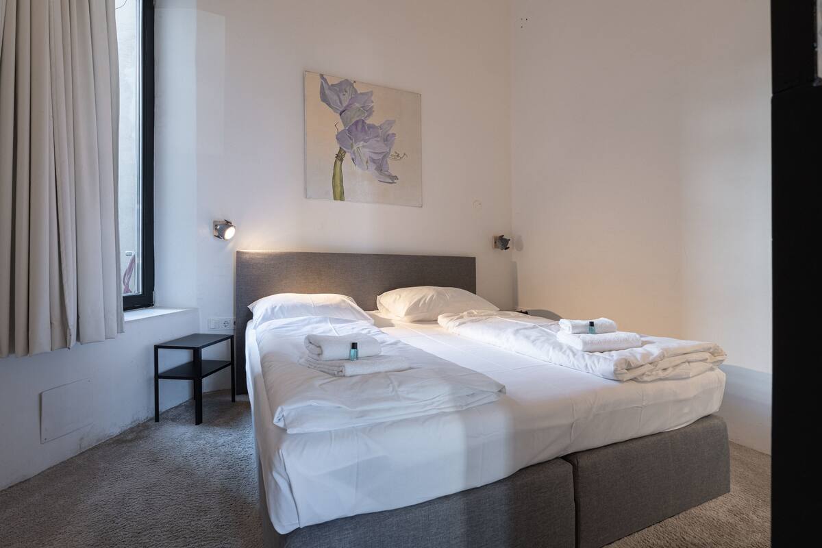 A neatly made bed with white linens, a bedside table, a wall-mounted light, and a window with curtains.