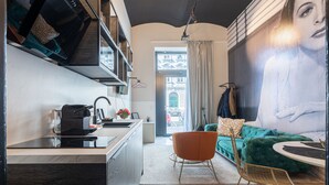A modern kitchen with a black countertop, a coffee machine, and a hanging rack. There is a teal sofa with a gold chair in the living area, and a large painting on the wall. - easy-flat Augarten - Apartments near Danube and Metro (Vienna)
