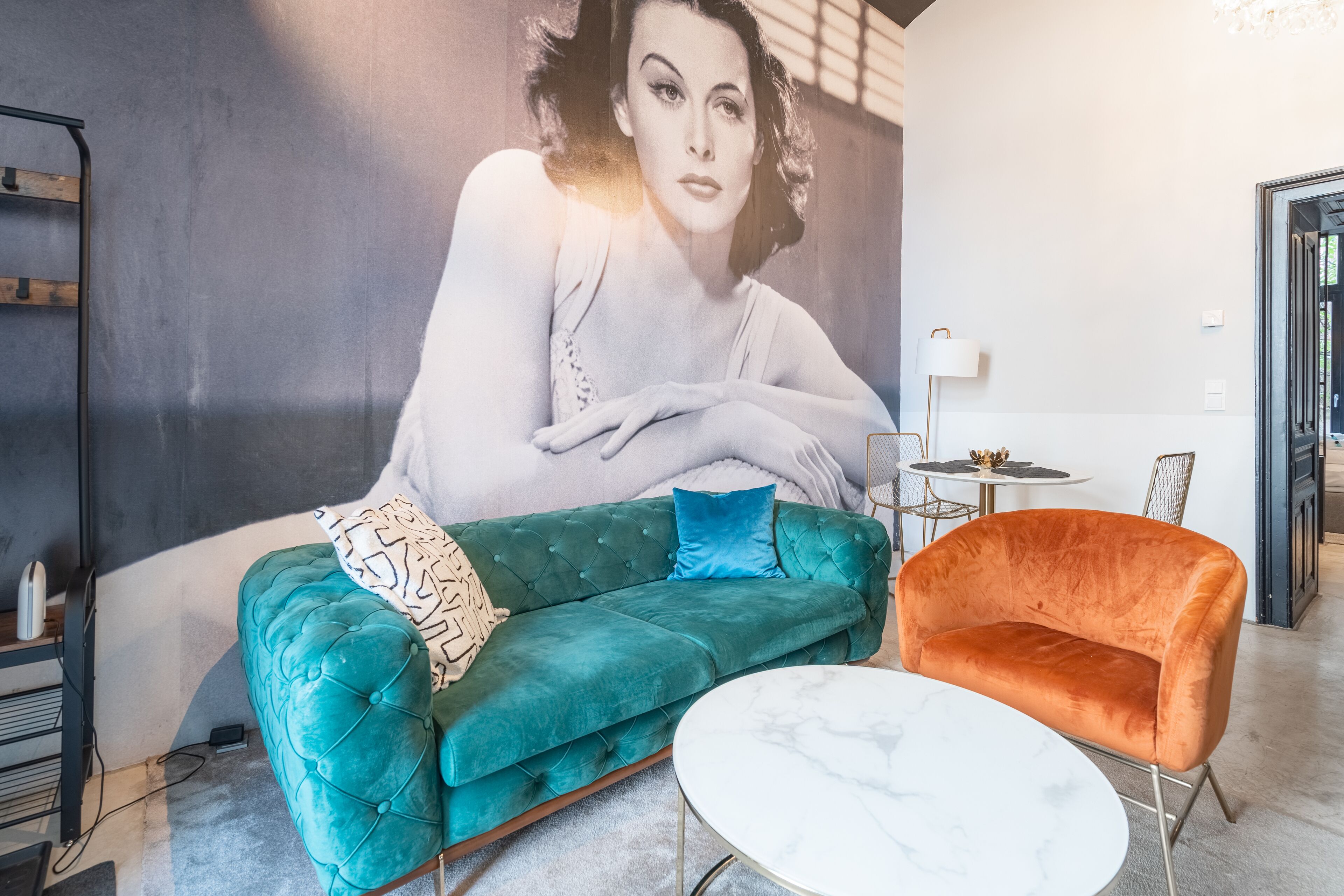 Ein blaugrünes Sofa mit einem blauen Kissen, ein orangefarbener Sessel und ein weißer Couchtisch in einem modernen Raum mit einer großen Wandmalerei.