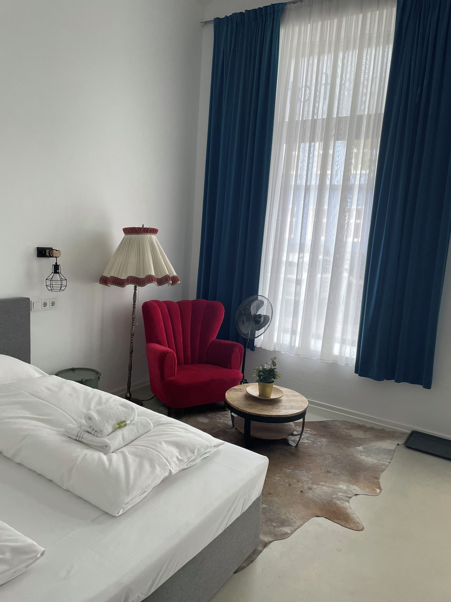 Habitación de hotel con cama, un sillón rojo, una mesita redonda con una planta, un ventilador de pie y un amplio ventanal con cortinas azules.