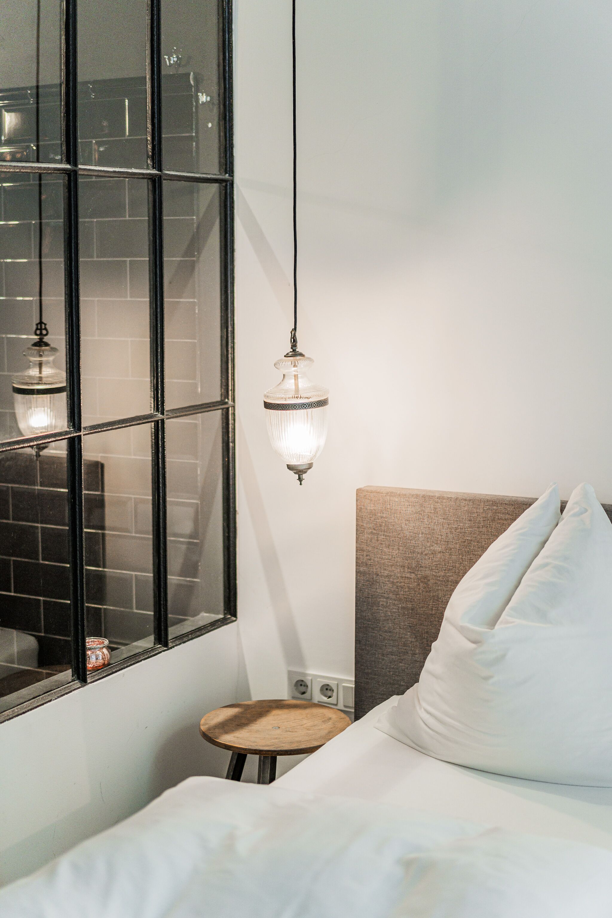 A hotel room with a bed, a small wooden stool, a window with a view of a brick wall, and a hanging light fixture.