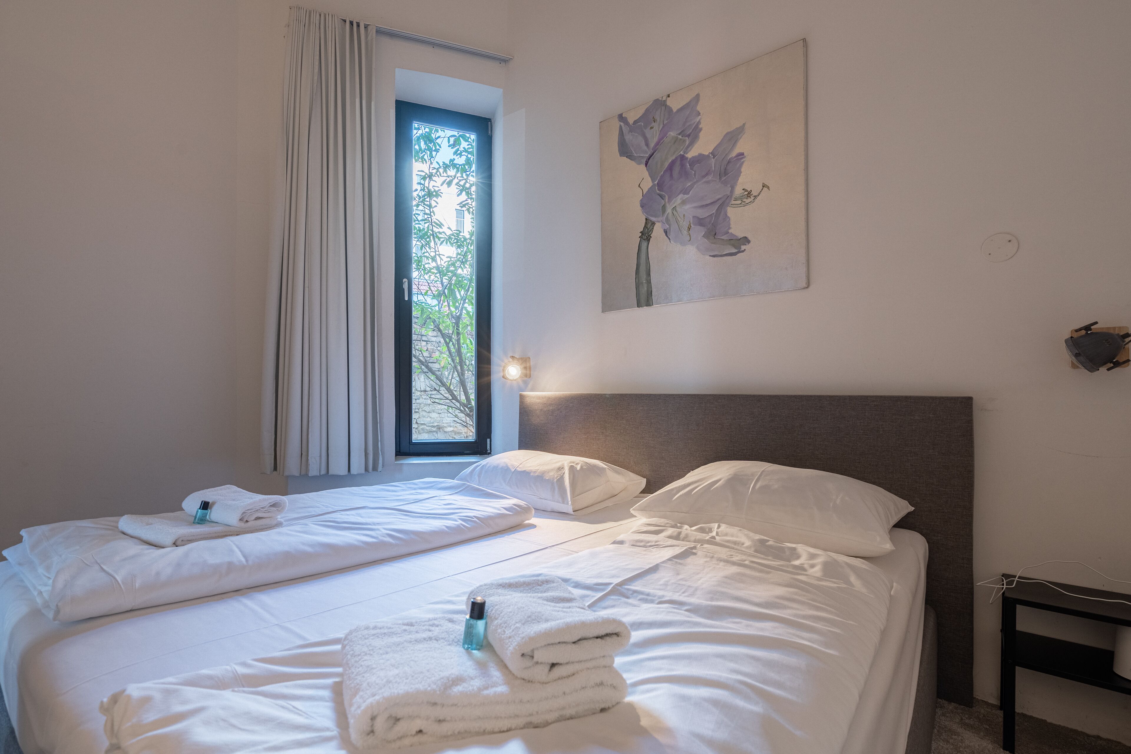 A hotel room with two beds, white linens, a painting of flowers, a window with curtains, and a bedside table with a lamp.