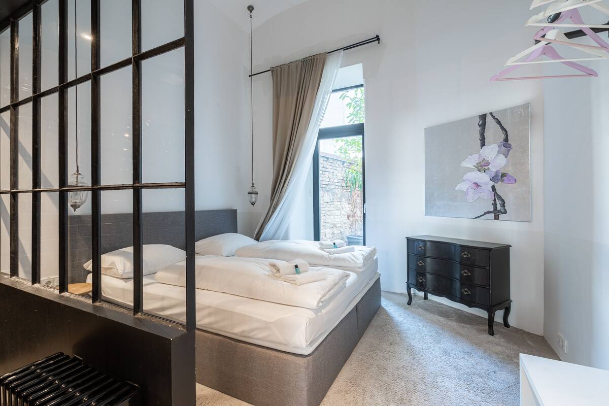 A modern bedroom with a large window, a bed with white linens, a dark dresser, and a wall-mounted artwork.