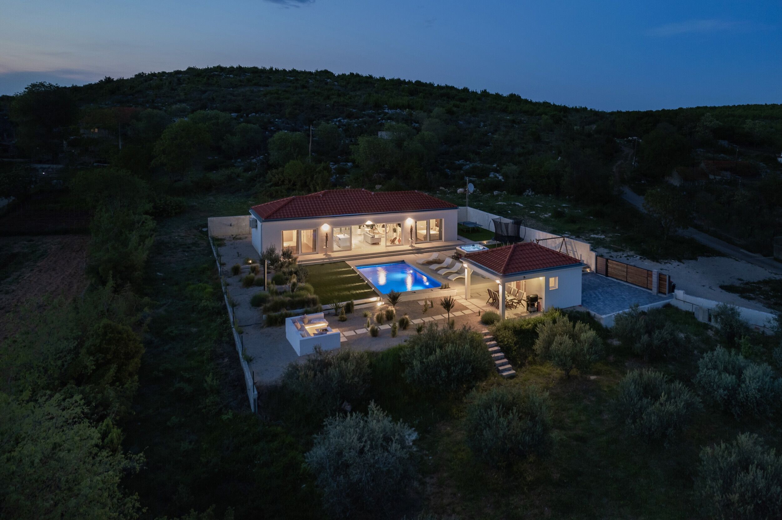 A modern two-story house with a swimming pool, surrounded by greenery and hills.