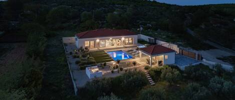 A modern two-story house with a swimming pool, surrounded by greenery and hills.