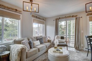 Un salon spacieux avec un canapé beige, un fauteuil blanc et une table basse. De grandes fenêtres offrent une vue sur l’extérieur.