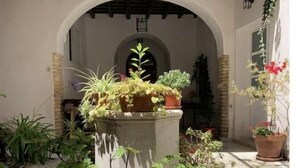 A courtyard with a stone fountain surrounded by potted plants and a white wall with a wooden balcony above.