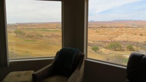 Une pièce dotée de deux grandes fenêtres donnant sur un paysage aride et dégagé, avec une végétation clairsemée et des collines à l’horizon.