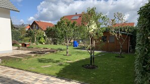 Un jardin à l’arrière d’une maison, avec une terrasse dallée, un petit arbre et un abri de jardin en bois.