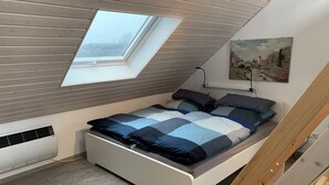 Une chambre de style loft moderne, avec un plafond en pente, une lucarne, un lit recouvert d’une courtepointe bleue et grise, et un tableau accroché au mur.