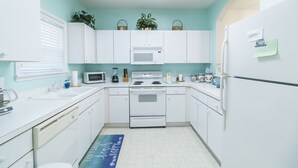 A compact kitchen with white cabinets, a microwave, stove, and a refrigerator. There are two potted plants on the countertop and a blue mat on the floor. - Beautiful Golf Villa, 2.7 Miles To Beach + Free Attraction Tickets! (North Myrtle Beach)