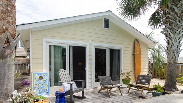 Une petite maison de plain-pied avec un porche abrité, deux portes-fenêtres coulissantes et une planche de surf appuyée contre le mur.