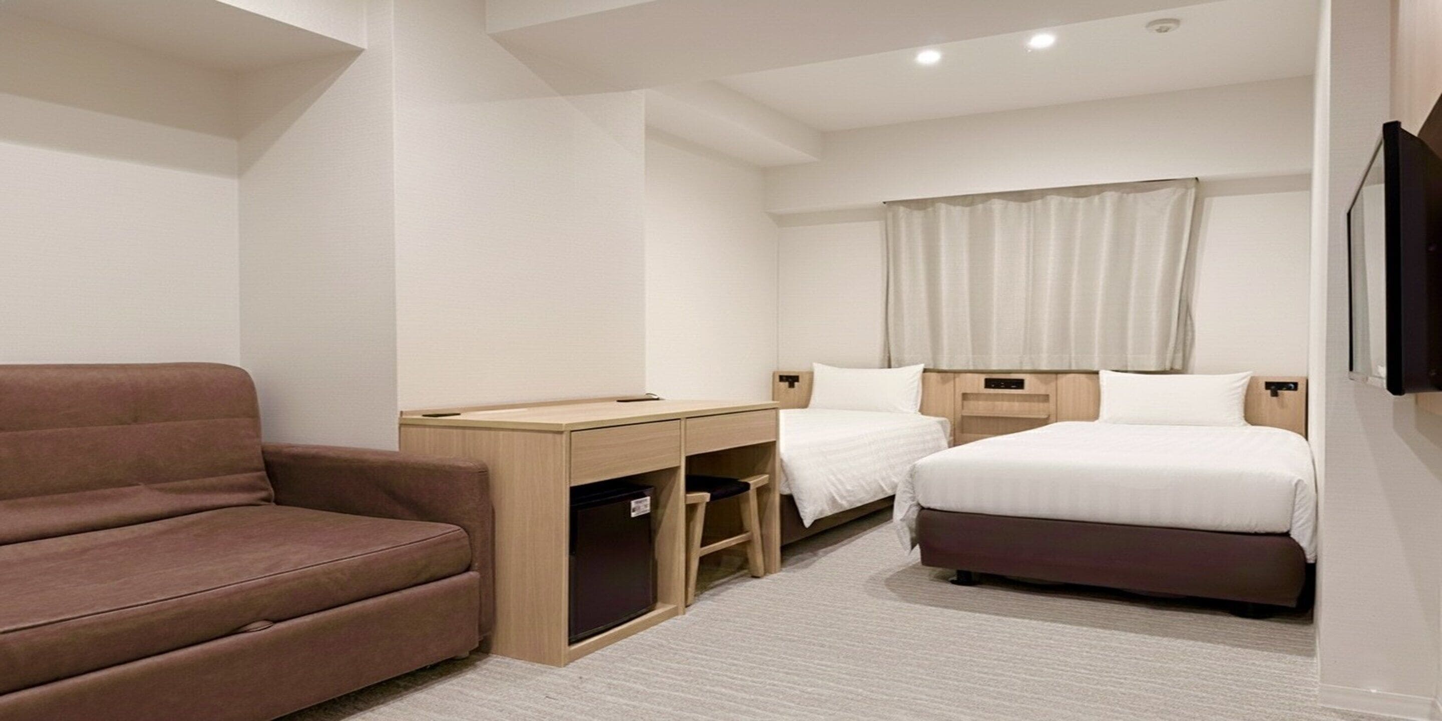 Ett hotellrum med en brun soffa, ett skrivbord med en dator, två sängar och en tv som är monterad på väggen.
