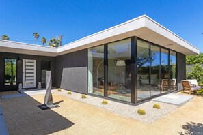 Une maison moderne avec de grandes fenêtres en verre, un patio avec des chaises et un jardin de cactus.
