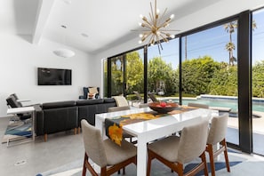 A modern living room with a dining area, large windows, a pool, and a black leather sofa.