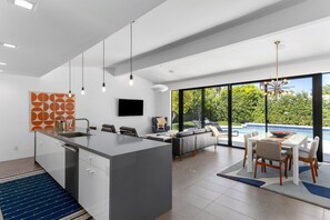 A modern kitchen with a central island, a dining area with a table and chairs, a sofa, and a large window overlooking a pool and greenery.