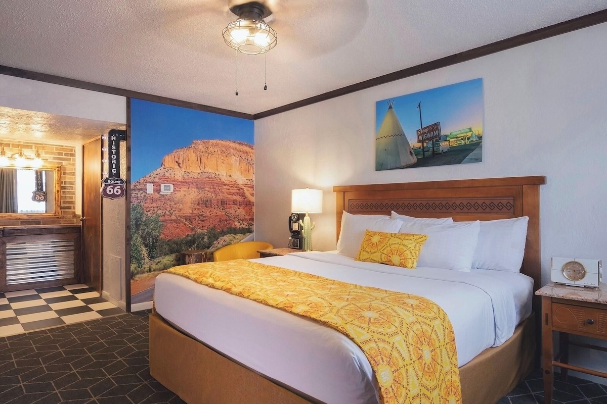 Habitación de hotel con cama, mesita de noche, lámpara, un cuadro de un paisaje desértico y un cartel en la pared.
