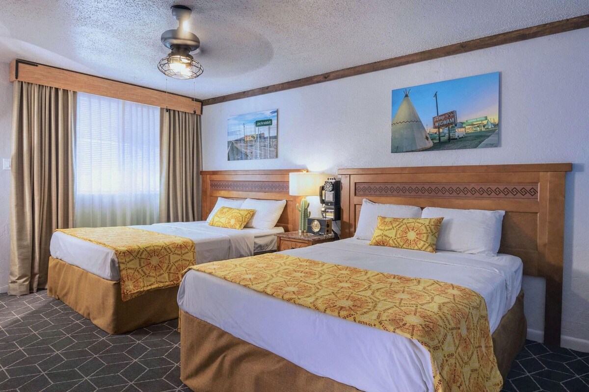 Habitación de hotel con dos camas, una mesita de noche con lámpara, ventilador de techo y un cuadro de un tipi.