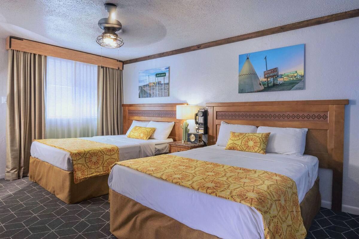 A hotel room with two beds, a nightstand with a lamp, a ceiling fan, and a painting of a teepee.