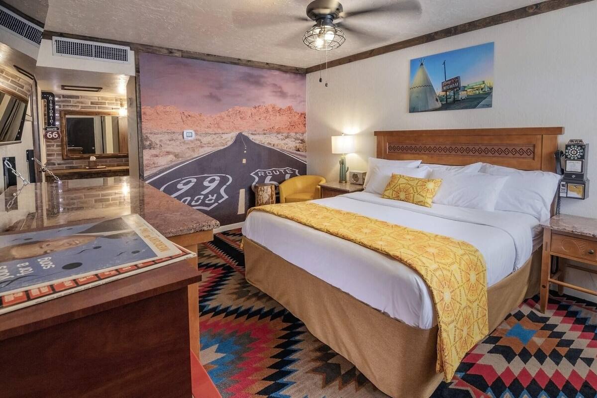 A hotel room with a bed, a desk, a mirror, a wall mural, a ceiling fan, and a nightstand.