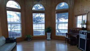 A spacious room with wooden walls, large arched windows, a piano, a couch, and a small table with a lamp. - Marina Pension (Ansan)